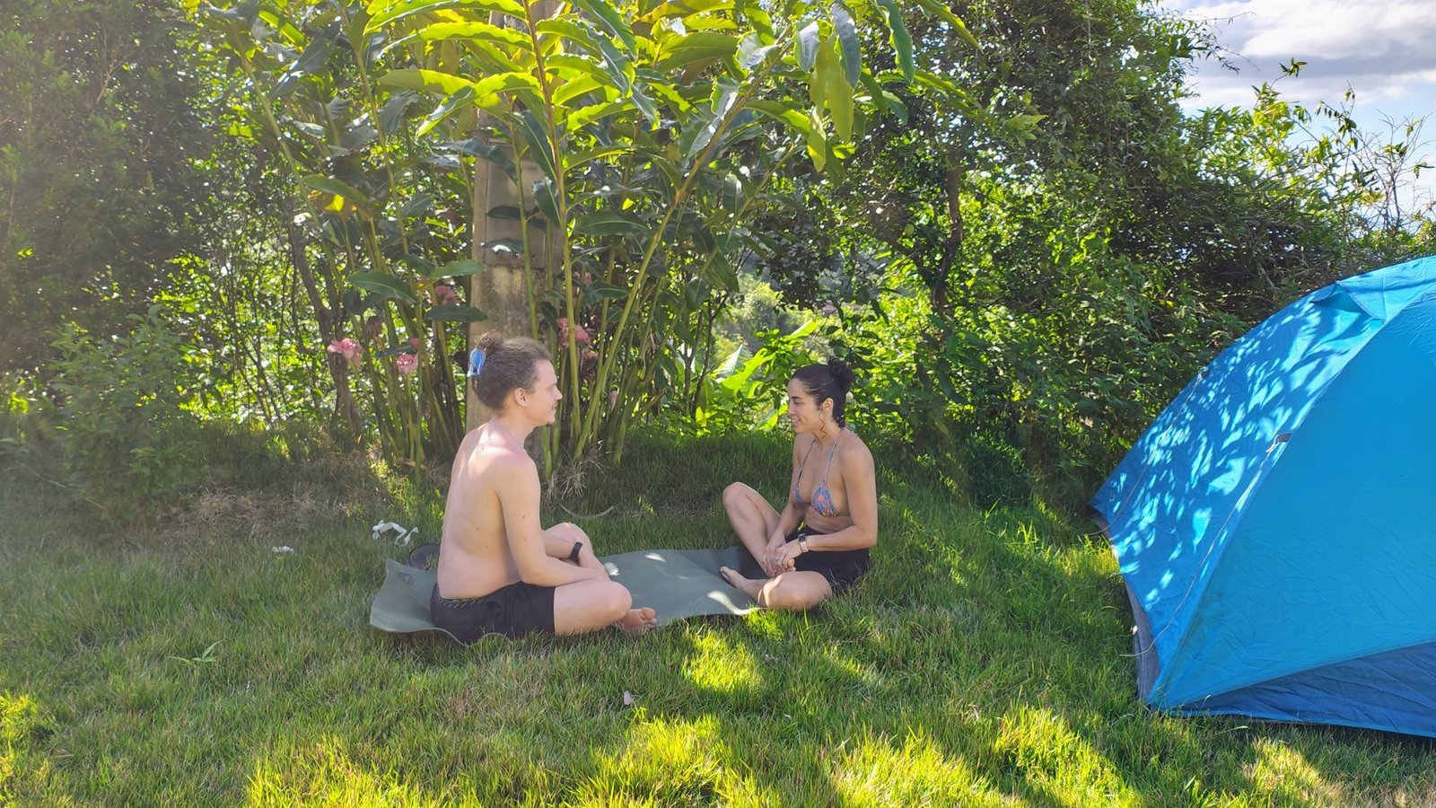 Duas pessoas conversando sentadas na grama, entre árvores e uma barraca